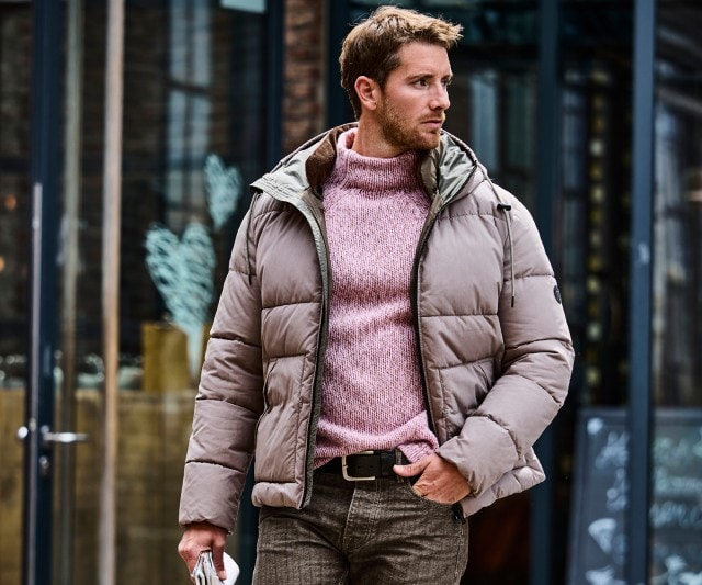 Ein Mann trägt eine taupefarbene Steppjacke über einem rosa-melierten Strickpullover mit Rollkragen, kombiniert mit einer braunen Hose und schwarzem Gürtel.