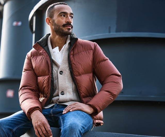 Ein Mann bekleidet mit einer weinroten Steppjacke sitzt auf einem Hocker und blickt in die Ferne.