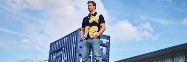 Ein Mann bekleidet mit einer Strickjacke und einer hellen Jeans steht auf einem Container und schaut in die Luft.