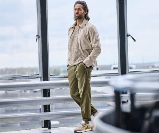 Ein Mann trägt eine beigefarbene Jacke, dazu eine Chinohose und Sneaker und schlendert durch ein Flughafengebäude.