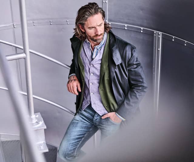 Ein Mann bekleidet mit einem grünen Sakko und einer blauen Jeans lehnt an einem Treppengeländer und blickt hinunter.