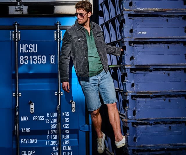 Ein Mann bekleidet mit einer Jeans Shorts einem grünen Polo und dunkler Jeansjacke kletter auf einen Seecontainer um sich einen besseren Überblick zu verschaffen