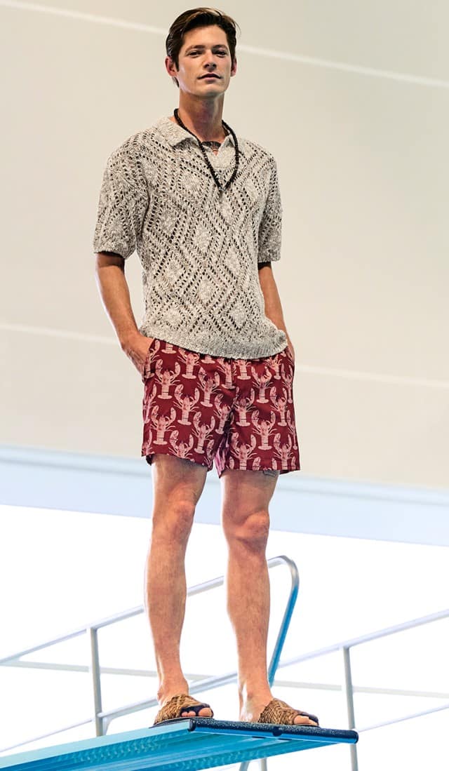 Ein Mann in einem hellen Poloshirt und rot-gemusterter Badehose steht auf eine Sprungbrett in einer Schwimmhalle.