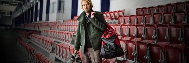 Das Bild zeigt einen Mann in einer leeren Eishalle, der Schlittschuhe und eine rote Sporttasche trägt.