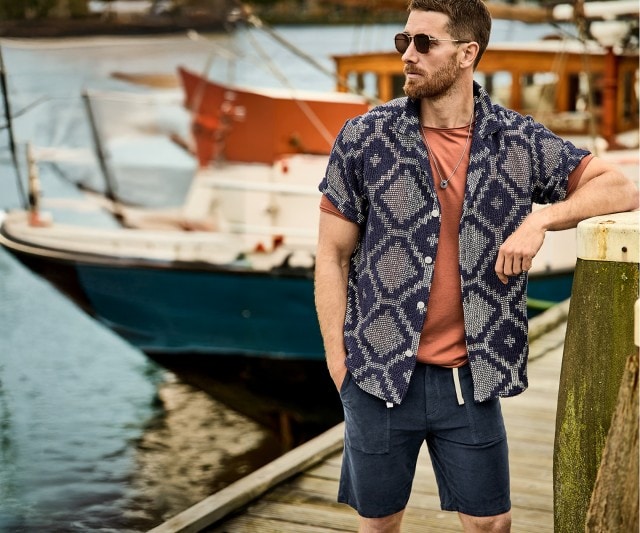 Mann trägt ein orangenes T-Shirt, darüber ein gemustertes Kurzarm-Polo und dunkelblaue Shorts