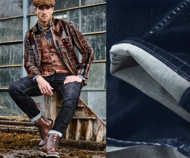 Auf dem Bild sitzt ein Mann lässig vor einem alten Industriegebäude, trägt eine braune Samtjacke mit Muster, eine Weste, ein Hemd, dunkle Jeans, braune Lederstiefel und eine Schirmmütze.