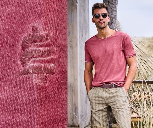 Ein Mann trägt ein rotes T-Shirt und gestreifte Hose, steht an einer Holzstruktur im Freien; daneben eine Nahaufnahme des Stoffes.