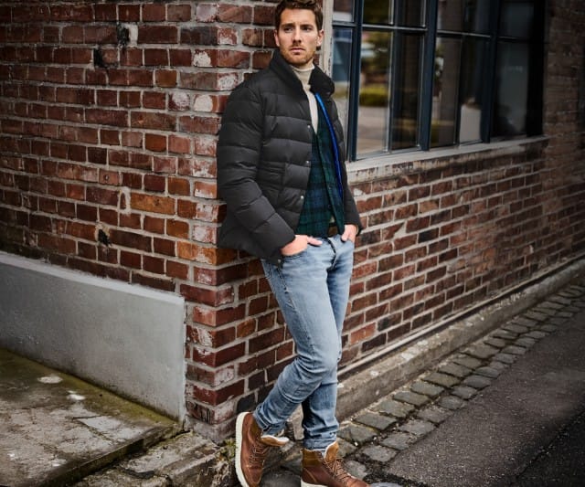 Auf dem Bild steht ein Mann lässig an einer Backsteinwand, trägt eine schwarze Steppjacke, einen Pullover, Jeans und braune Winterstiefel.
