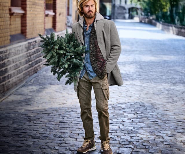 Auf dem Bild ist ein Mann zu sehen, der auf einer gepflasterten Straße geht und einen kleinen Tannenzweig trägt; er ist lässig gekleidet mit grauem Mantel, Jeanshemd, Strickjacke, Cargohose und braunen Stiefeln.
