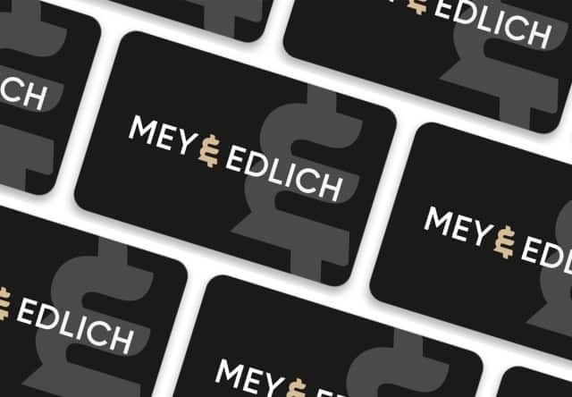 Auf dem Bild sind mehrere schwarze Karten mit dem weißen „MEY & EDLICH“-Logo und einem großen grauen grafischen Element im Hintergrund diagonal angeordnet.