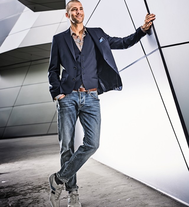 Mann mit blauem Sakko und Jeans steht an einem Außengebäude und lächelt