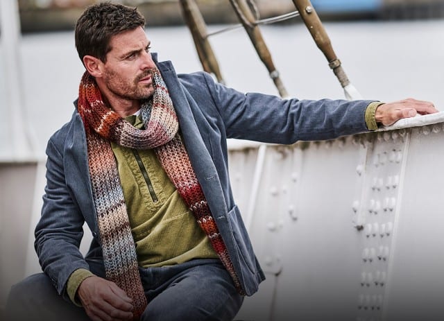 Ein Mann sitzt an Bord eines Schiffs, trägt eine blaue Jacke, ein olivgrünes Oberteil und einen mehrfarbigen Strickschal, während er nachdenklich zur Seite blickt.