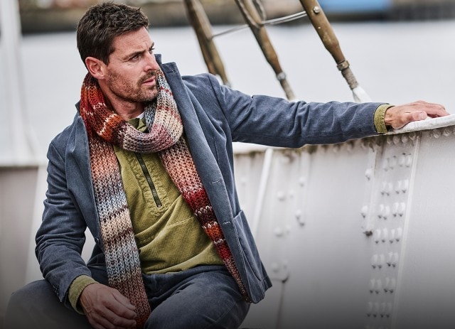 Ein Mann sitzt lässig auf einem Schiff und trägt einen blau-grauen Mantel mit grünem Shirt und einem bunt gestrickten Schal, was ihm einen stilvoll-herbstlichen Look verleiht.