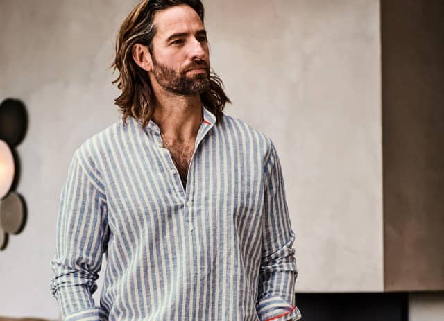 Ein Mann mit schulterlangem Haar und Bart trägt ein hellblau-weiß gestreiftes Hemd mit offenem Kragen und blickt ruhig zur Seite.