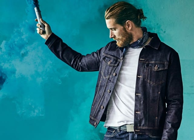 Ein Mann mit zurückgebundenen Haaren und Bart trägt eine dunkle Jeansjacke über einem weißen T-Shirt, hält eine Rauchfackel in der Hand und steht vor einer türkisblauen Rauchkulisse.