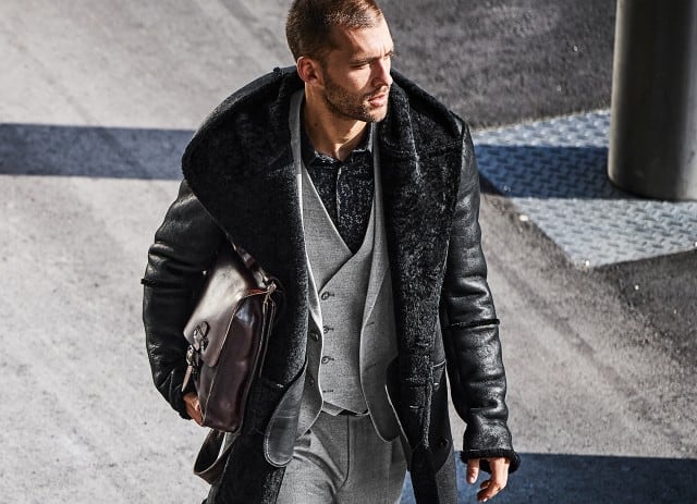 Ein Mann geht im Sonnenlicht über eine Straße, trägt einen eleganten grauen Anzug mit schwarzer Lammfelljacke und hält eine braune Ledertasche in der Hand.
