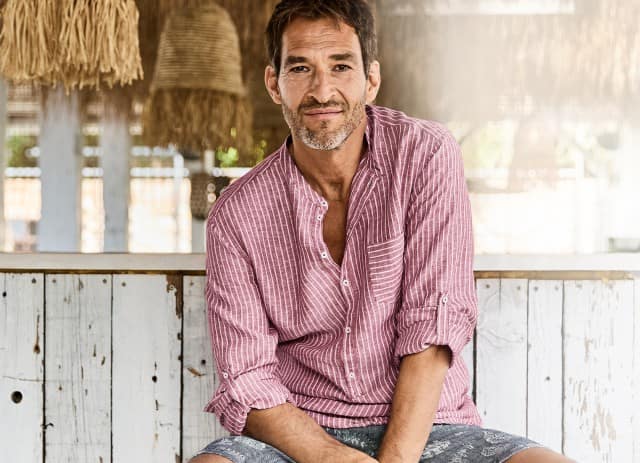 Ein Mann sitzt entspannt in einer hellen, sommertypischen Umgebung, trägt ein rosafarbenes, gestreiftes Hemd mit hochgekrempelten Ärmeln und lächelt leicht in die Kamera.