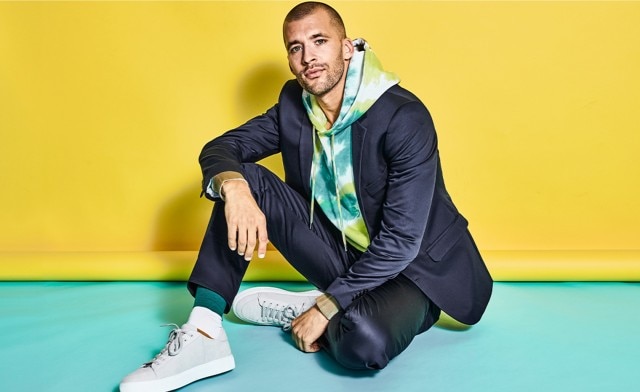 Sitzender Mann vor einem gelben Hintergrund in einem blauen Anzug und bunten Hoodie.
