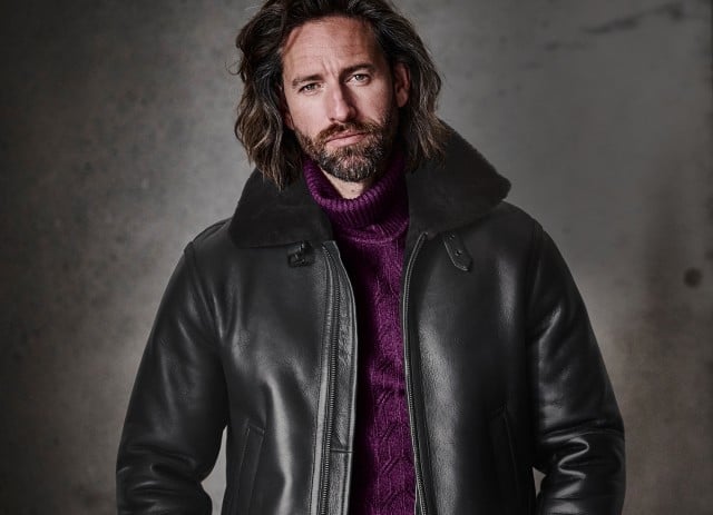 Ein Mann mit mittellangem Haar und Bart trägt einen lila Strickrollkragenpullover unter einer schwarzen Lederjacke mit breitem Kragen und blickt ruhig in die Kamera.