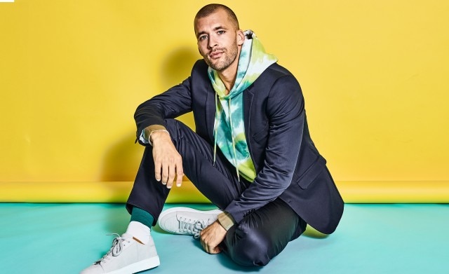 Sitzender Mann vor einem gelben Hintergrund in einem blauen Anzug und bunten Hoodie