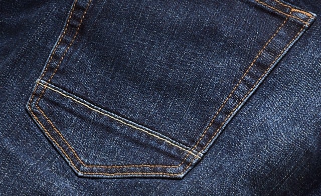 Das Bild zeigt eine Nahaufnahme von dunkelblauem Denimstoff mit einer aufgesetzten Gesäßtasche und kontrastierenden, orangefarbenen Nähten.