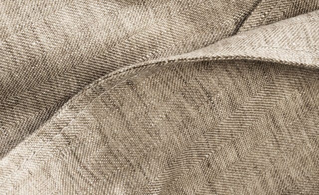 Das Bild zeigt eine Nahaufnahme eines beigefarbenen Leinens mit sichtbarer Webstruktur und feinem, leicht unregelmäßigem Fadenverlauf.