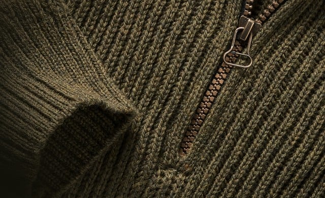 Das Bild zeigt eine Nahaufnahme eines grob gestrickten Merinowollpullovers in Olivgrün mit einem metallenen Reißverschluss.