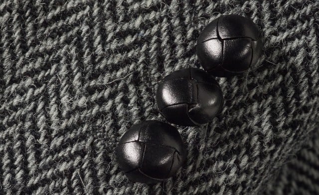 Auf dem Bild ist eine Nahaufnahme eines grau‑schwarzen Fischgrätstoffs mit drei dunklen, geflochten wirkenden Knöpfen zu sehen.