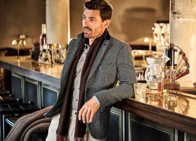 Ein Mann lehnt lässig an einer Bar, trägt einen grauen Fischgrät‑Blazer und einen dunklen Schal, während vor ihm ein Glas und eine Flasche Whisky stehen.