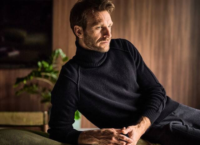 Ein Mann sitzt lässig auf einem Schiff und trägt einen blau-grauen Mantel mit grünem Shirt und einem bunt gestrickten Schal, was ihm einen stilvoll-herbstlichen Look verleiht.