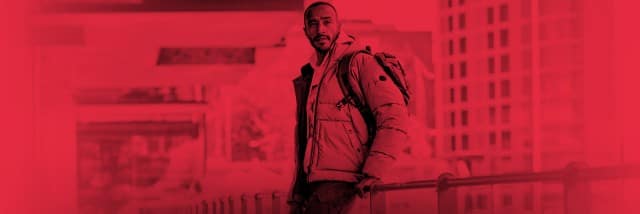 Ein Mann steht draußen in einer urbanen Umgebung an einem Geländer, trägt eine dicke Winterjacke und einen Rucksack und blickt nach in die Kamera.