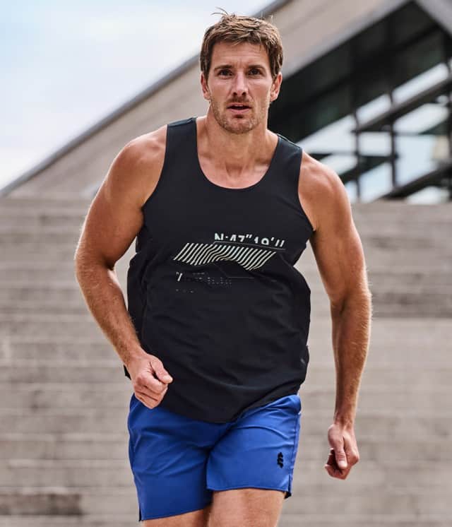 Ein sportlich gekleideter Mann läuft energisch eine Treppe hinunter, trägt ein schwarzes Tanktop und blaue Shorts und wirkt dabei konzentriert und entschlossen.