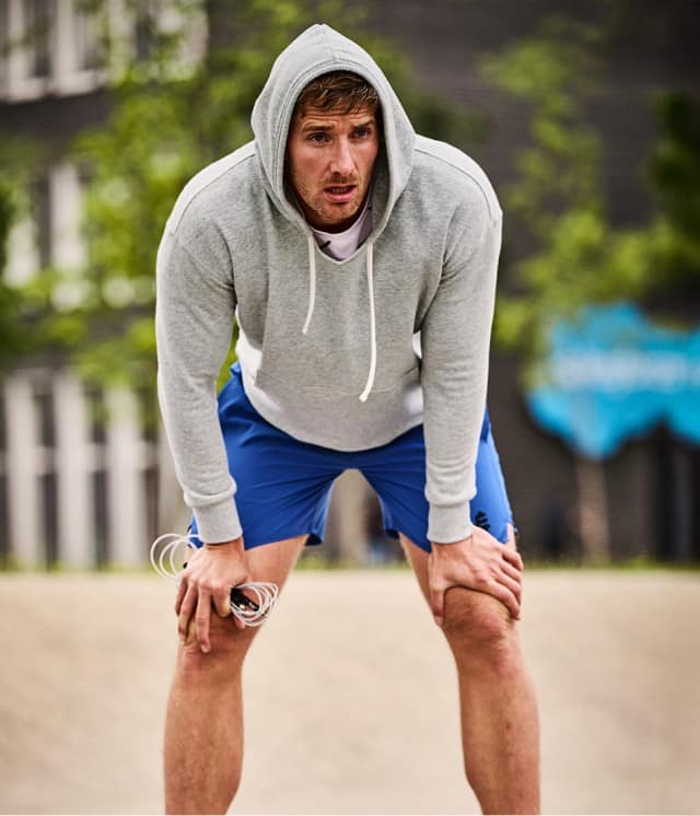 Ein erschöpft wirkender Mann in grauem Hoodie und blauen Shorts beugt sich nach vorne und stützt die Hände auf die Knie, während er draußen eine kurze Pause vom Training macht.