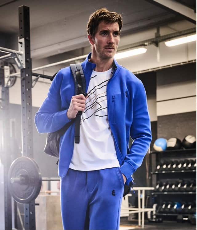 Ein Mann in einem blauen Trainingsanzug und weißem T‑Shirt steht im Fitnessstudio und trägt einen Rucksack über der Schulter.