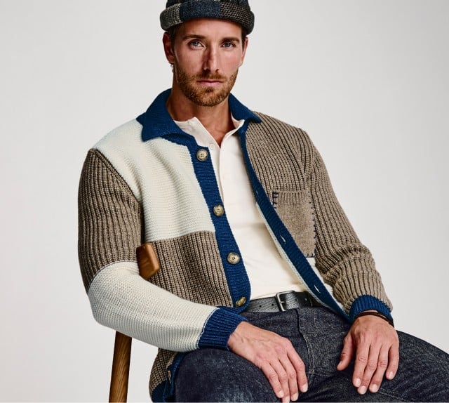 Ein Mann sitzt auf einem Stuhl und trägt eine mehrfarbige Strickjacke, ein helles Shirt, dunkle Jeans und eine passende Mütze vor hellem Hintergrund.