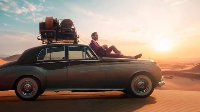 Ein Mann in elegantem Anzug sitzt entspannt auf der Motorhaube eines Oldtimers, der inmitten einer Wüstenlandschaft bei Sonnenuntergang steht, mit Gepäck auf dem Dach.