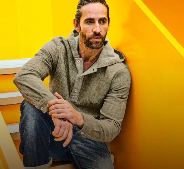 Der Mann trägt ein graues Kapuzenshirt mit Knopfleiste und eine dunkelblaue Jeans.