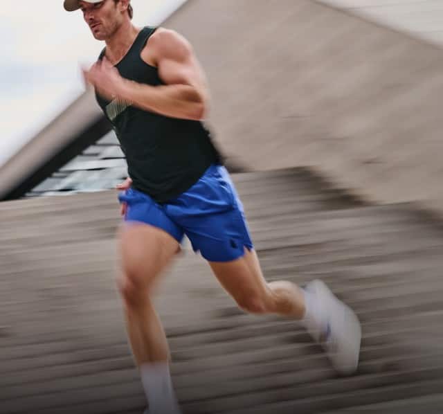 Ein Mann joggt dynamisch eine Treppe hinunter und trägt dabei ein dunkles Tanktop, blaue Shorts, eine Kappe und Sportschuhe.