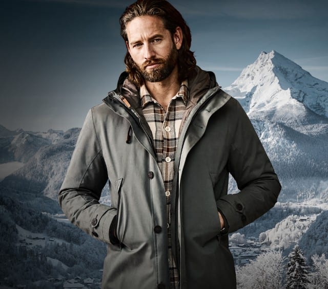 Das Bild zeigt einen Mann in einer grauen Winterjacke über einem karierten Hemd vor schneebedeckter Bergkulisse.