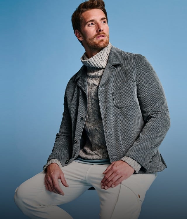 Das Bild zeigt einen Mann in winterlicher Kleidung – bestehend aus Strickrollkragen, grauer Jacke und heller Hose – vor blauem Hintergrund.