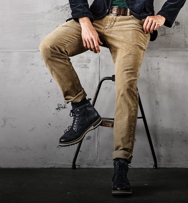 Das Bild zeigt einen Mann, der beige Cordhosen und schwarze Boots trägt und lässig auf einem Hocker sitzt.