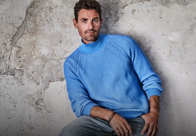 Das Bild zeigt einen Mann, der einen hellblauen Strickpullover und eine hellgraue Jeans trägt.