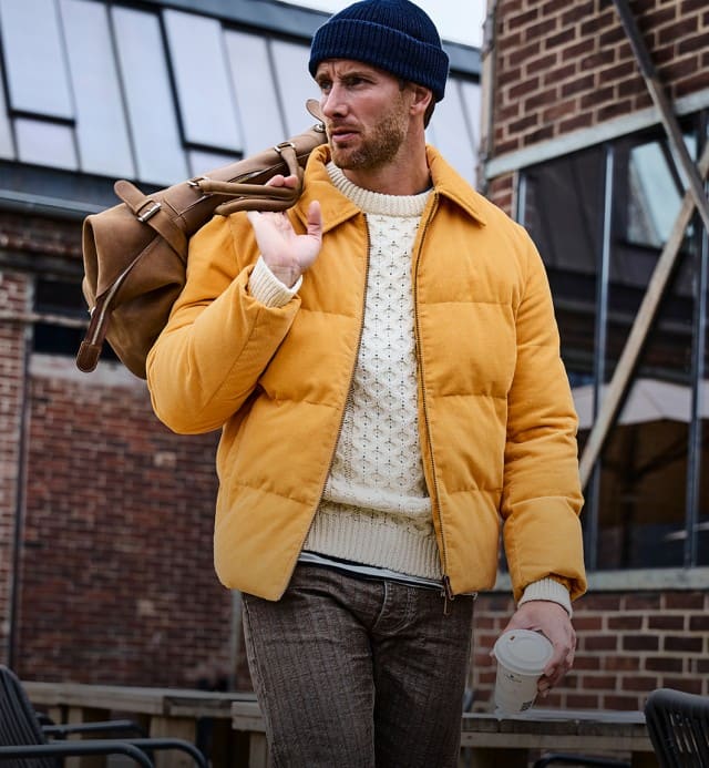 Ein Mann trägt eine gelbe Steppjacke über einem cremefarbenen Strickpullover, kombiniert mit einer dunklen Hose und einer dunkelblauen Mütze.