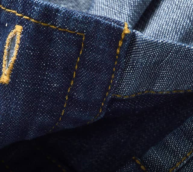 Zu sehen ist eine Nahaufnahme eines Jeansstoffs mit gelber Kontrastnaht, Knopfloch und Knopfdetail.