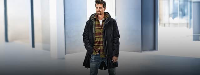 Ein Mann trägt einen langen dunkelblauen Mantel über einem gestreiften Hoodie in Grün und Braun, kombiniert mit einer hellen Shirtlage und einer verwaschenen blauen Jeans.