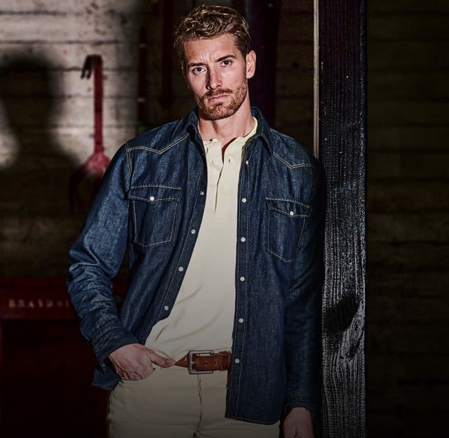 Der Mann trägt ein dunkelblaues Jeanshemd über einem hellen Polohemd sowie eine helle Hose mit braunem Gürtel.