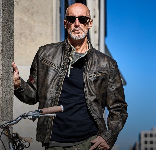 Ein Mann trägt eine braune Lederjacke über einem dunklen Shirt und einer aufgeknöpften Henley‑Shirt‑Leiste, kombiniert mit einer Sonnenbrille.