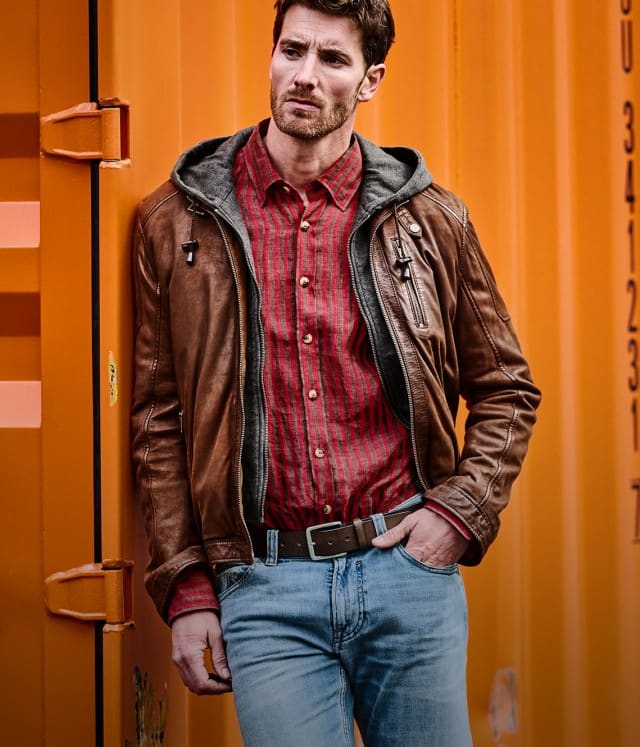 Ein Mann bekleidet mit einer braunen Lederjacke, einem roten Hemd und einer hellblauen Jeans lehnt lässig an einem orangefarbenen Container.