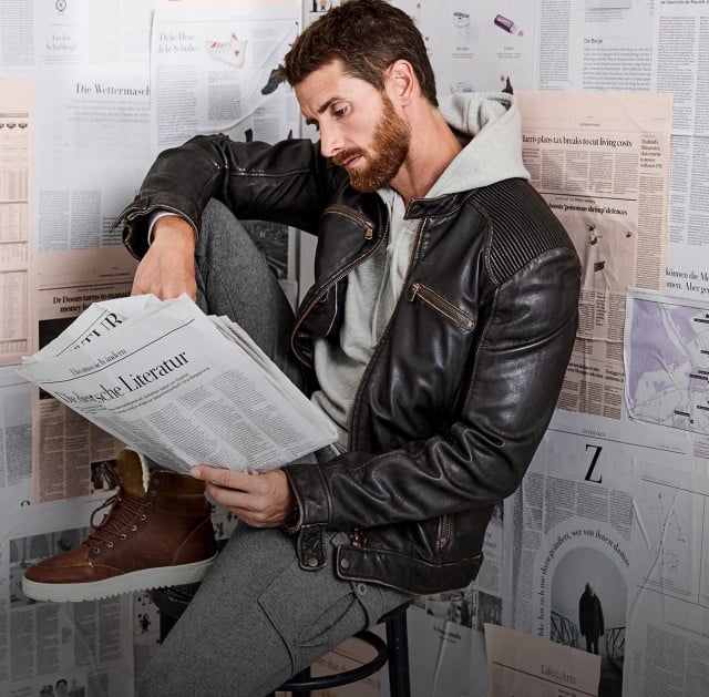 Der Mann trägt eine schwarze Lederjacke über einem grauen Hoodie, dazu eine graue Jogpant und braune Schnürstiefel.