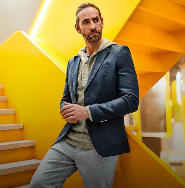 Der Mann trägt einen dunkelblauen Blazer über einem hellgrünen Kapuzenshirt sowie eine graue Hose.